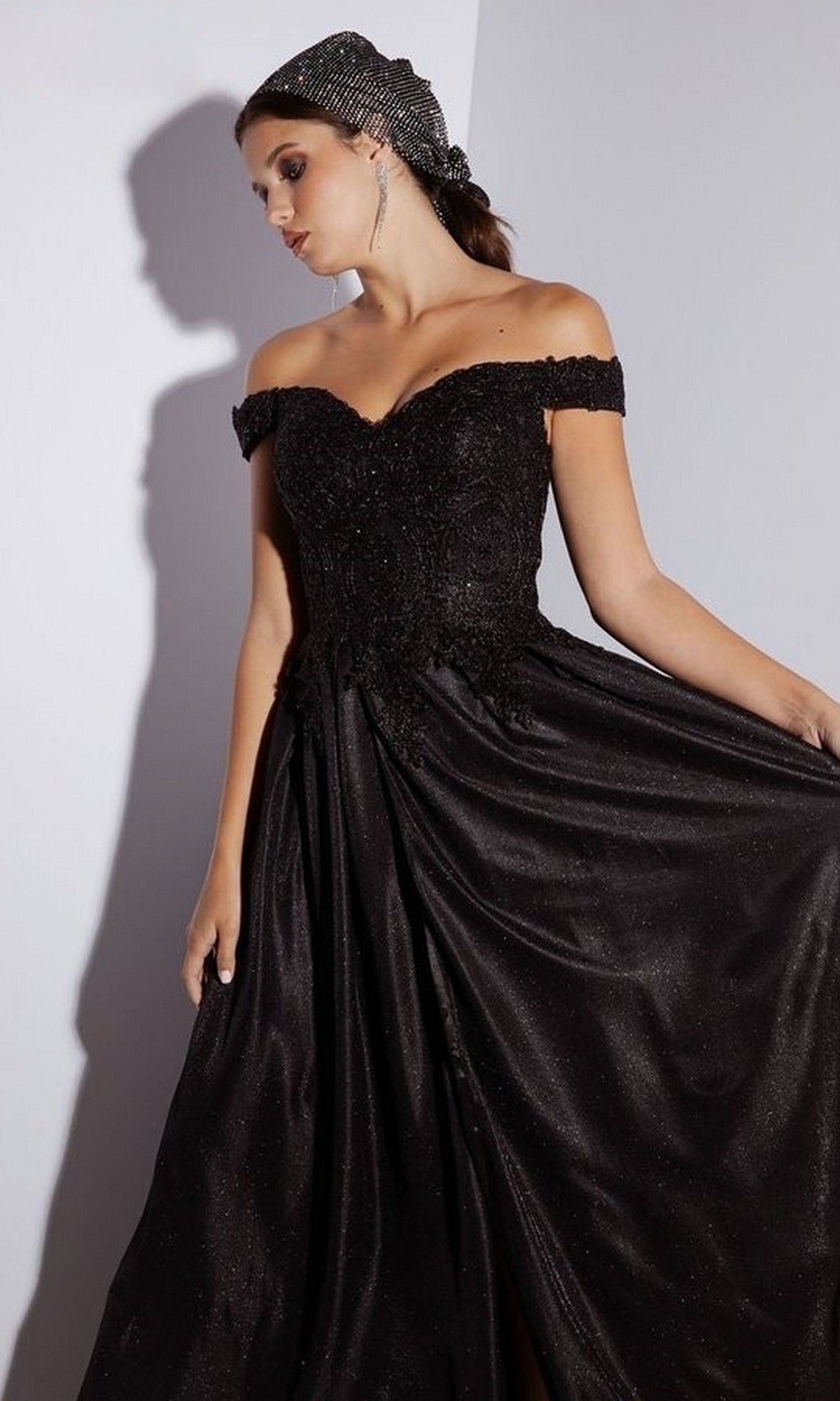 A woman stands against a white wall in the black Eureka 9806 long prom dress, featuring an off-the-shoulder beaded bodice, lace applique details, and a matching headscarf.